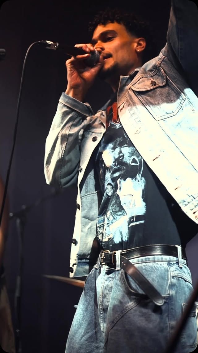 OSK on stage at night in a light denim jacket over a vintage rap tee, singing into a microphone.
