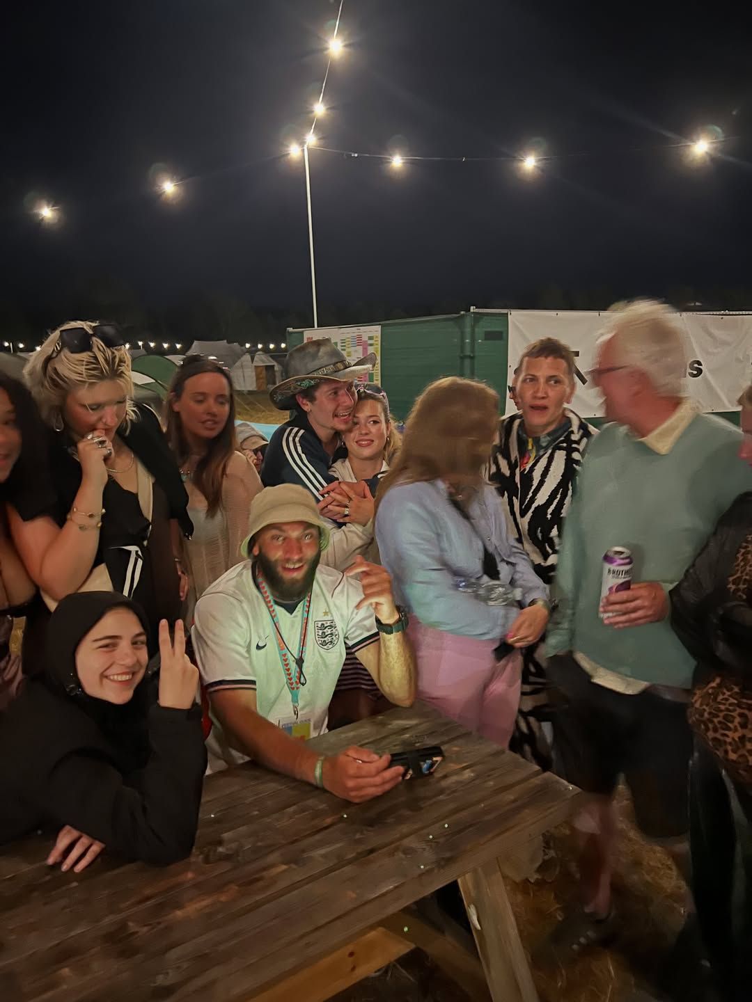 A crowd of friends packed around a picnic table under festoon lights at night, OSK in the middle.