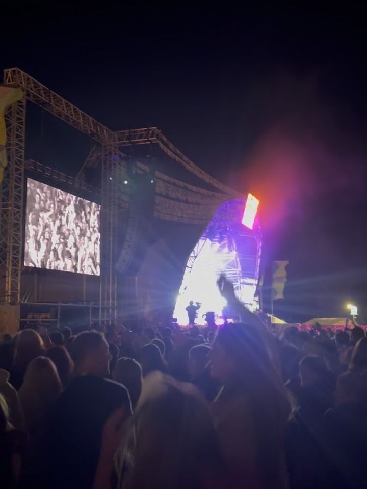 A festival night stage glowing purple and white over a dark crowd, a big screen to one side.