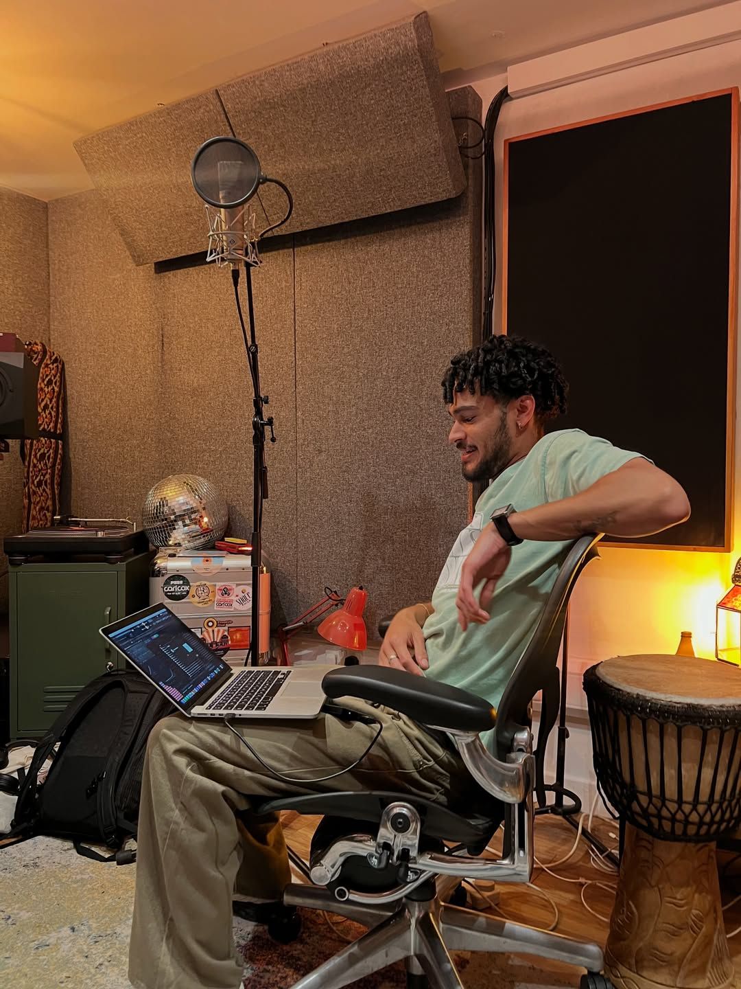 OSK leaning back in a desk chair in a small home studio, laptop on his knee, a microphone and a djembe beside him.