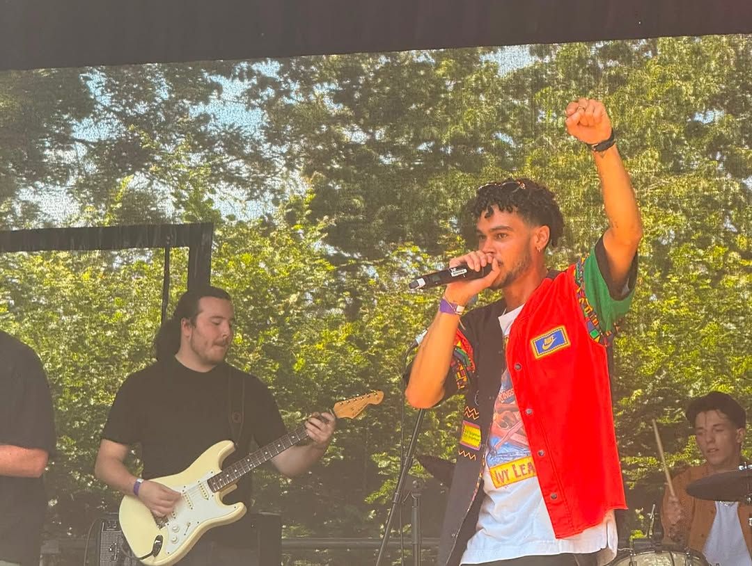 OSK in a red, green and yellow jacket, arm raised, fronting the full band on a sunlit outdoor stage under the trees, a guitarist beside him.
