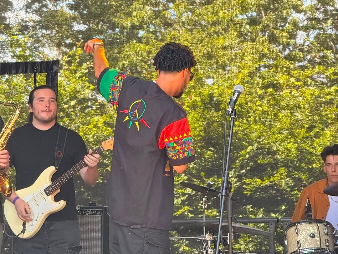 OSK in a pan-African colours jacket on the outdoor stage with a guitarist in a patterned shirt beside him and the drummer behind.