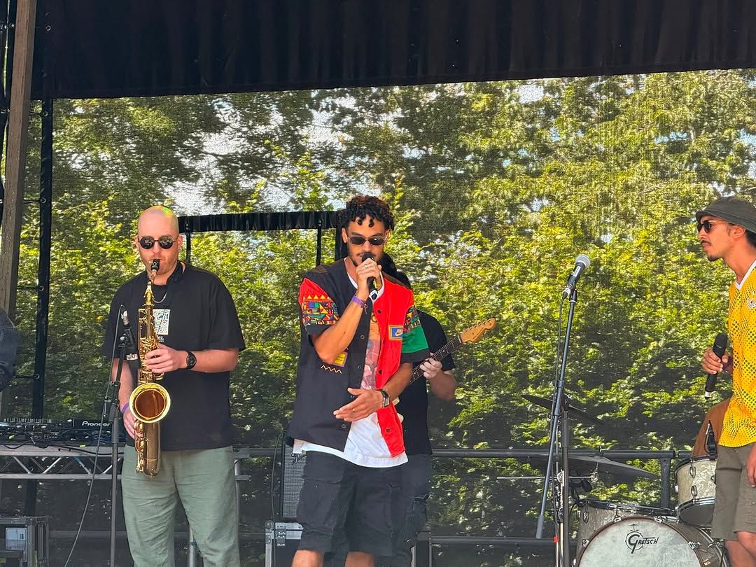 The band mid-set on the outdoor stage: a saxophonist and OSK at the front, drums and guitar behind.