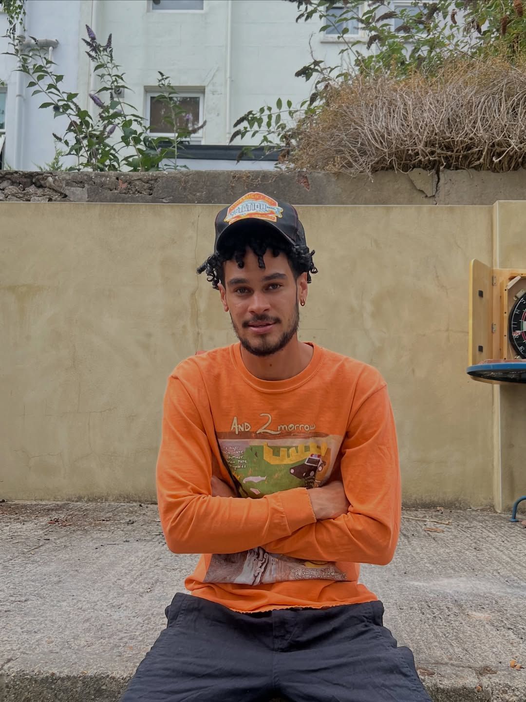 OSK sitting cross-armed on a low wall in a sunny Brighton backyard, wearing a bright orange graphic tee and a cap.